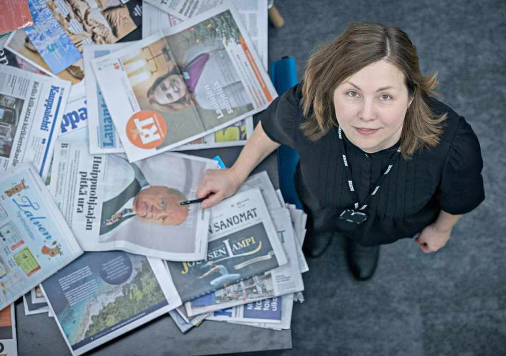 Toimittaja Silja Aitoaho katsoo ylös kameraan,. vieressään sanomalehtiä.