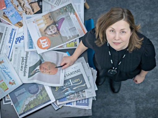 Toimittaja Silja Aitoaho katsoo ylös kameraan,. vieressään sanomalehtiä.